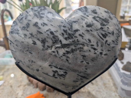 Large polished heart of milky quartz with dense forest-green actinolite inclusions on a black stand, front view.