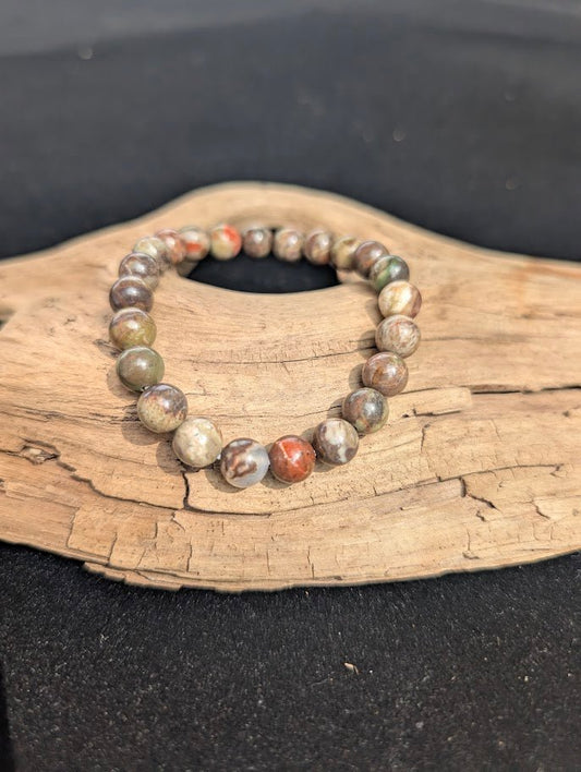An 8mm Money Agate beaded bracelet resting on natural driftwood, displaying earthy red, green, and grey chalcedony colours.