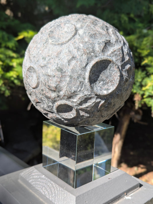 A 12 kg Yooperlite carved moon sphere in natural sunlight, showing its textured grey syenite matrix resting on a clear cubic stand.