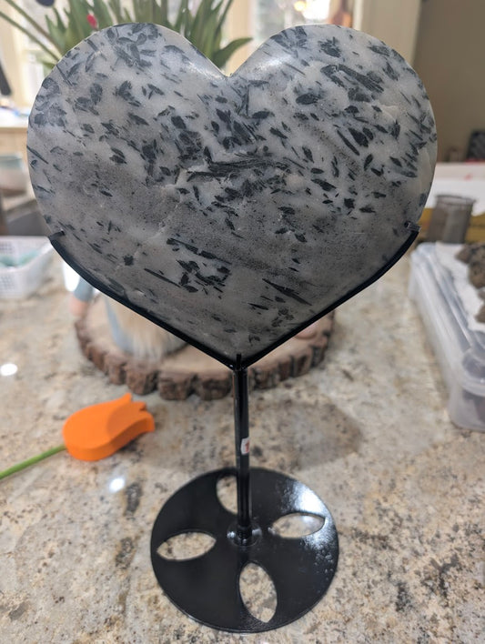 Large polished heart of milky quartz with dense forest-green actinolite needle inclusions on a black metal stand.