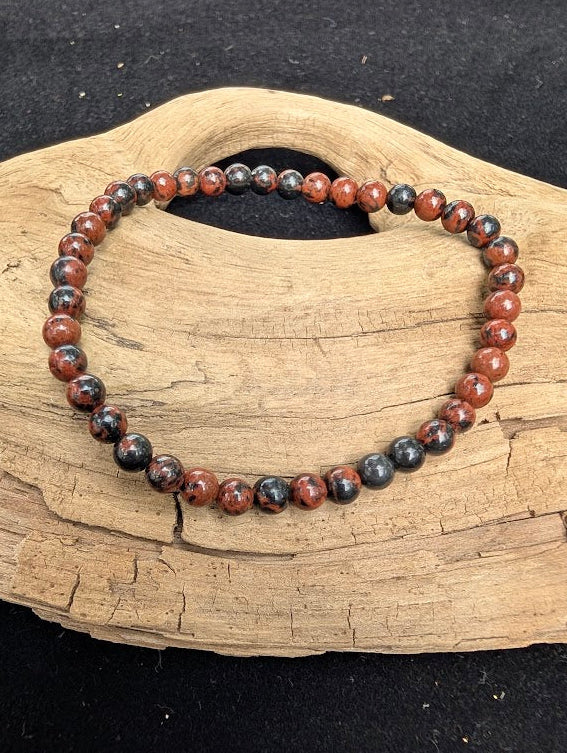 4mm polished Mahogany Obsidian beaded stretch bracelet resting on a driftwood display, showing swirled black and red tones.