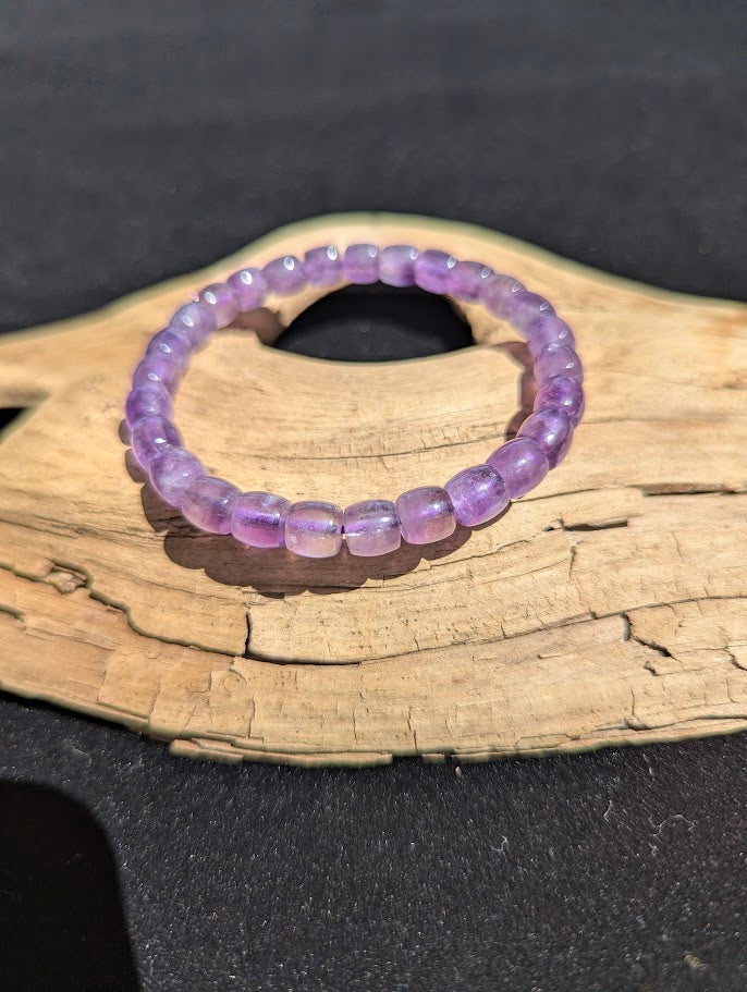 Polished 7mm Amethyst barrel bead stretch bracelet resting on a driftwood display, showing translucent violet and lavender tones strung on a purple cord.