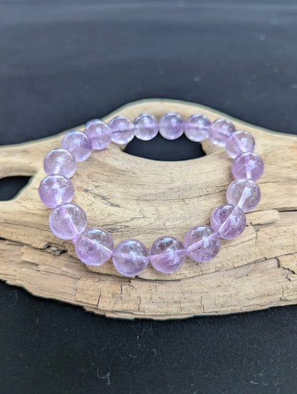 12mm High Quality Brazilian Amethyst beaded stretch bracelet resting on a driftwood display, showing translucent lavender spherical quartz beads on a clear cord.