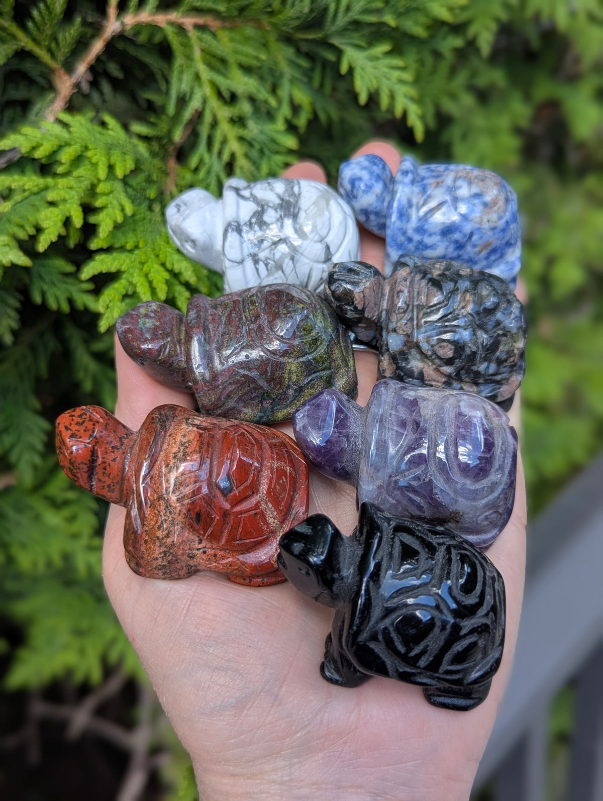 Hand holding multiple geometric turtle carvings carved from various minerals including howlite, sodalite, amethyst, obsidian, and unakite.