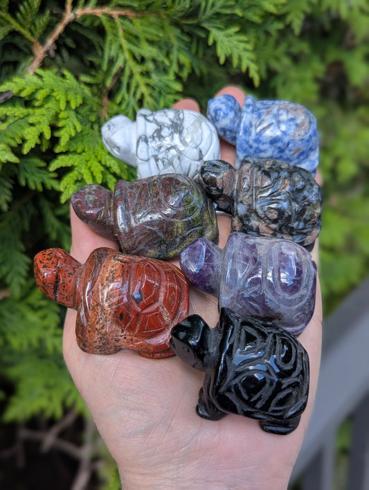 Hand holding multiple geometric turtle carvings carved from various minerals including howlite, sodalite, amethyst, obsidian, and unakite.