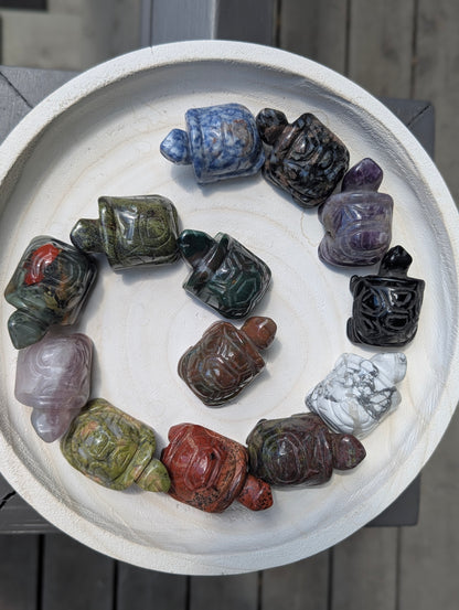 A variety of geometric mineral turtle carvings arranged in a spiral on a white tray, showcasing different stone types like jasper, amethyst, and sodalite.