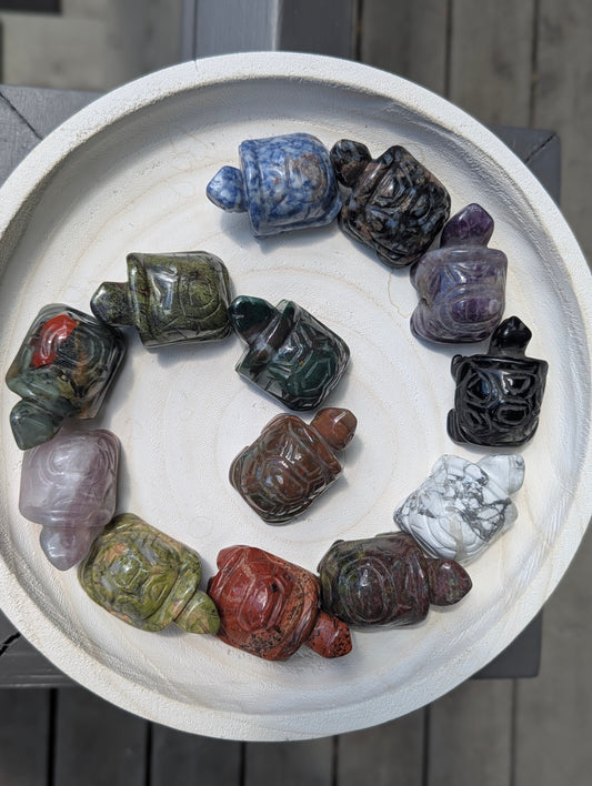 A variety of geometric mineral turtle carvings arranged in a spiral on a white tray, showcasing different stone types like jasper, amethyst, and sodalite.