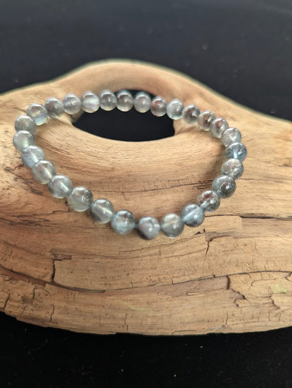 Intuitively selected 7mm AAA Grade Brazilian Girasol Black Devil Aquamarine beaded stretch bracelet displayed on natural driftwood, showcasing translucent stormy grey-blue beryl beads.