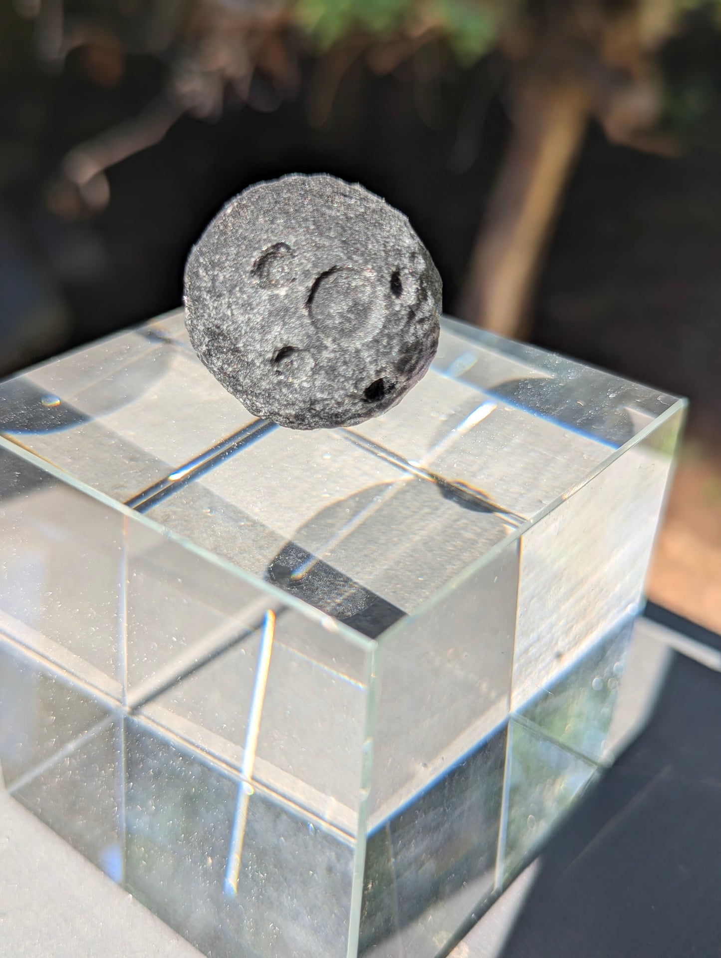 A 30 mm (1.18 in) Black Obsidian mini carved moon sphere resting on a clear cubic display stand in natural sunlight.