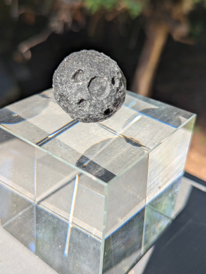 A 30 mm (1.18 in) Black Obsidian mini carved moon sphere resting on a clear cubic display stand in natural sunlight.