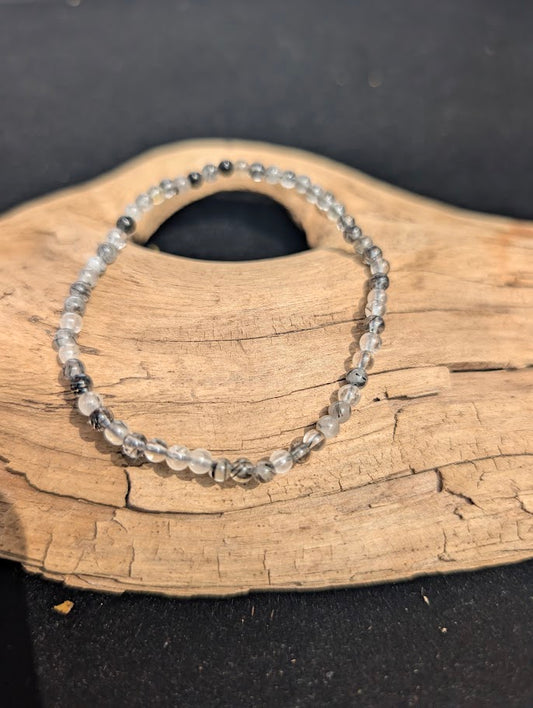 A 4mm Black Rutilated Quartz beaded bracelet resting on natural driftwood, displaying translucent quartz with black tourmaline needle inclusions.