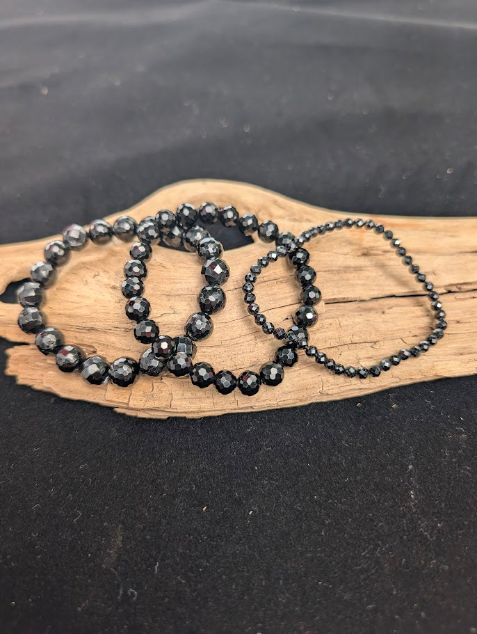 Three sizes of faceted Black Tourmaline beaded bracelets displayed together on driftwood.