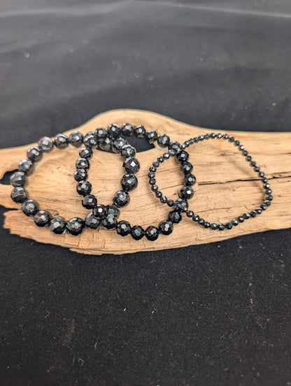 Three sizes of faceted Black Tourmaline beaded bracelets displayed together on driftwood.