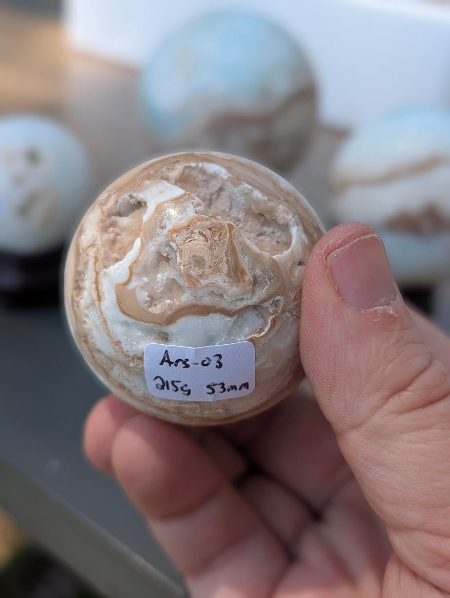 Base view of the 53mm Blue Aragonite sphere held in hand, showing a jagged vug and the 215g weight label for variant Ars-03.