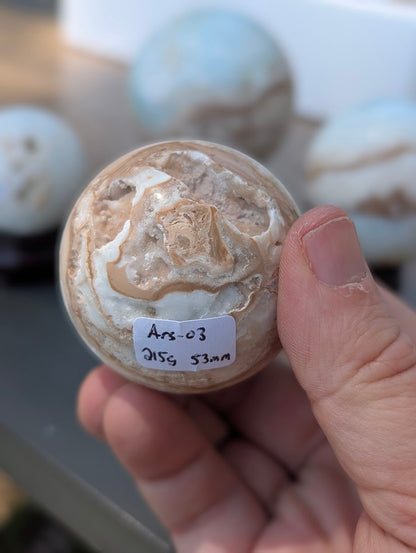 Base view of the 53mm Blue Aragonite sphere held in hand, showing a jagged vug and the 215g weight label for variant Ars-03.