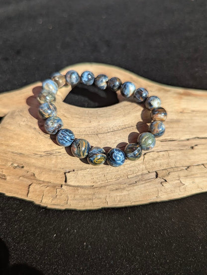 Intuitively selected 10mm AAA Grade Namibian Blue Pietersite beaded stretch bracelet resting on driftwood, showcasing high optical clarity and swirling fibers.