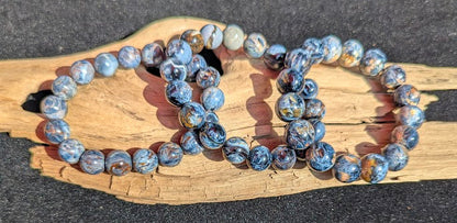 Suite of intuitively selected AAA Grade Namibian Blue Pietersite beaded stretch bracelets displayed together on driftwood, highlighting the multi-directional chatoyancy and brecciated matrix across different bead sizes.