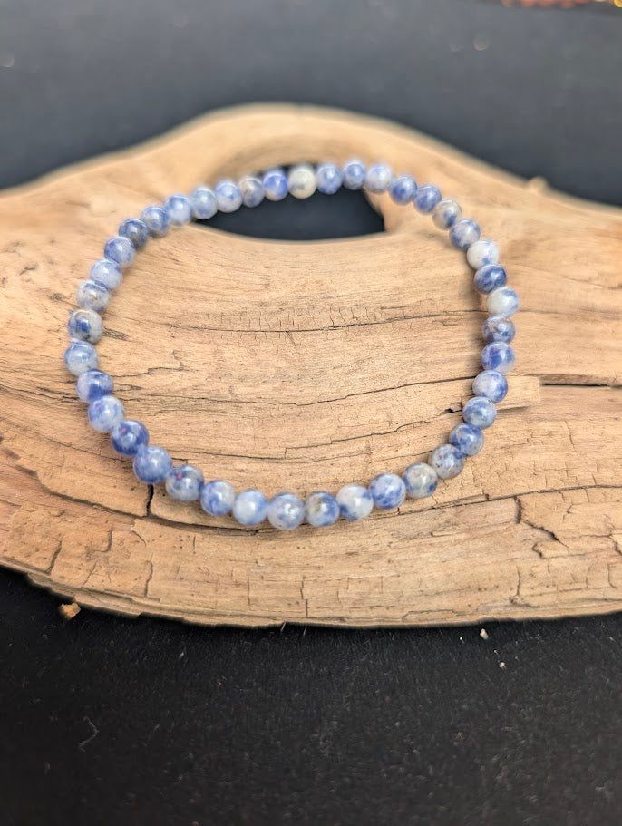 A 4mm Blue Point Jasper beaded bracelet resting on natural driftwood, displaying mottled denim blue and white colours.