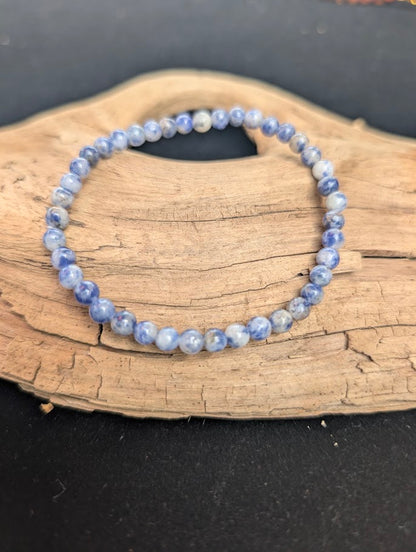 A 4mm Blue Point Jasper beaded bracelet resting on natural driftwood, displaying mottled denim blue and white colours.