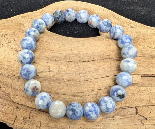 8mm faceted Blue Point Stone beaded stretch bracelet resting on a driftwood display, showing mottled denim blue and milky white Dumortierite Quartz tones.