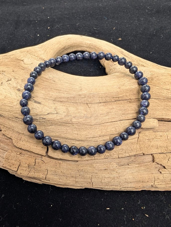4mm polished Blue Sandstone beaded stretch bracelet resting on a driftwood display, showing a dark blue matrix with sparkly inclusions.