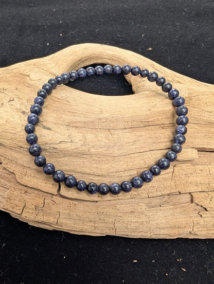 4mm polished Blue Sandstone beaded stretch bracelet resting on a driftwood display, showing a dark blue matrix with sparkly inclusions.