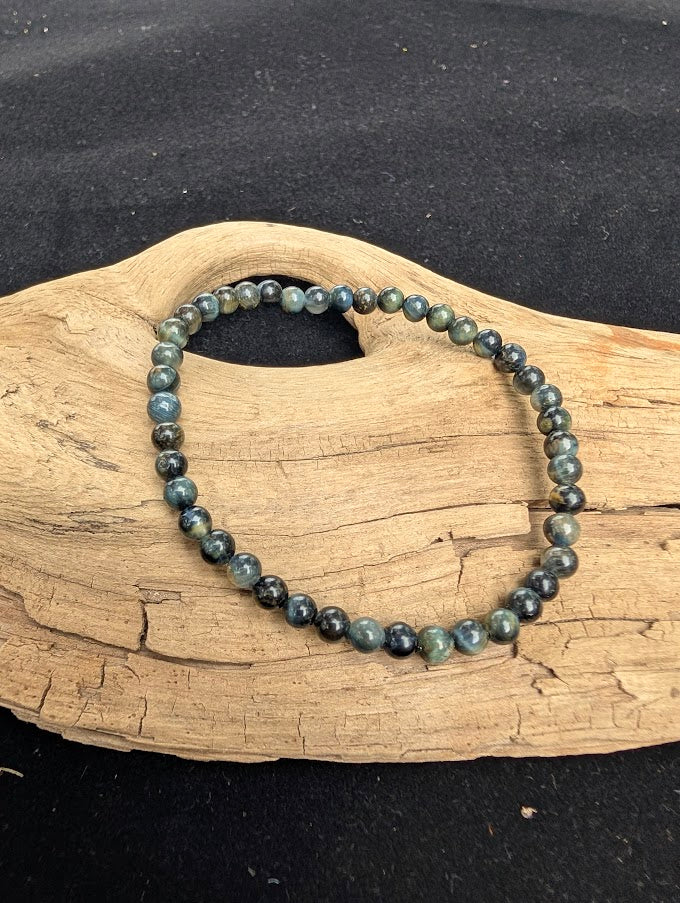 4mm polished Blue Tiger Eye beaded stretch bracelet resting on a driftwood display, showing dark blue-grey tones and natural chatoyancy.