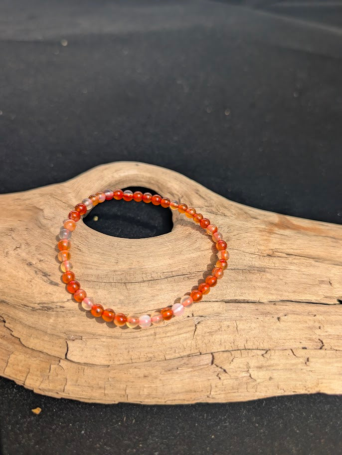 A 4mm Carnelian beaded bracelet resting on natural driftwood, displaying translucent peach and vibrant orange-red colours.