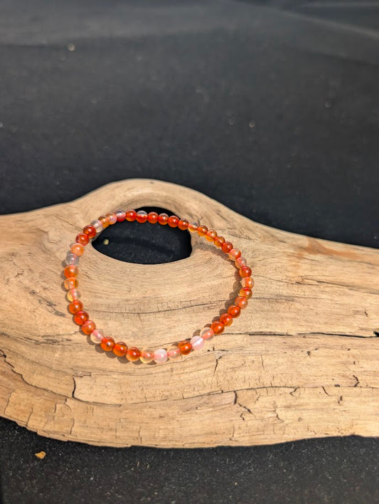 A 4mm Carnelian beaded bracelet resting on natural driftwood, displaying translucent peach and vibrant orange-red colours.