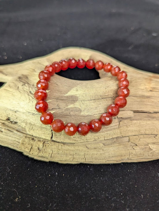 8mm faceted Carnelian beaded stretch bracelet resting on a driftwood display, showing vibrant orange and deep red translucent tones.