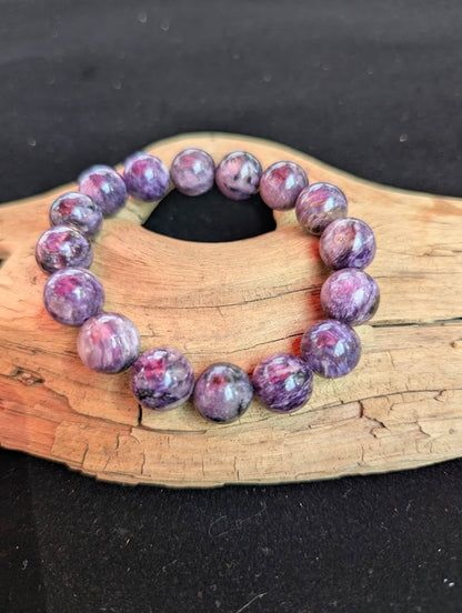 Intuitively selected 13mm AAA grade Siberian Charoite beaded stretch bracelet on a driftwood display, showing prominent reddish-pink inclusions.