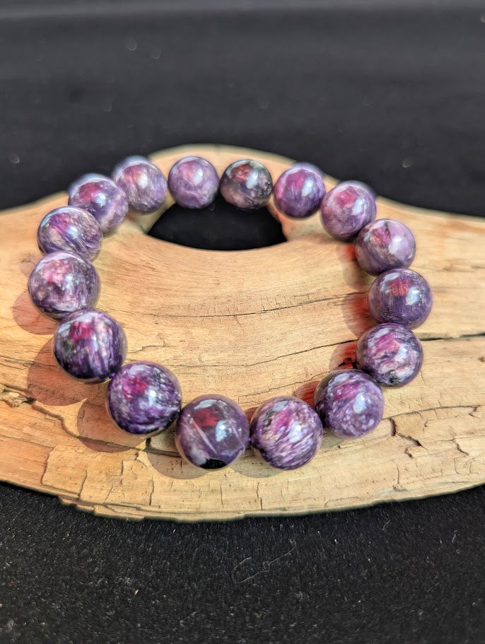 Intuitively selected 14mm AAA grade Siberian Charoite beaded stretch bracelet on a driftwood display, showing prominent reddish-pink inclusions.