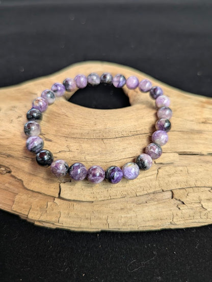 Intuitively selected 7mm AAA grade Siberian Charoite beaded stretch bracelet resting on a driftwood display, showcasing vibrant purple swirling patterns.