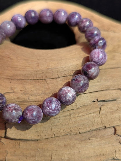 Macro photograph of a 9mm AAA Charoite beaded bracelet showing high vitreous lustre, purple cord detailing, and intricate mineral banding.