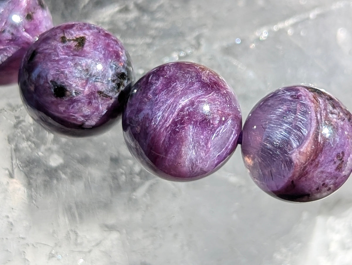 Macro close-up of three AAA Siberian Charoite beads showing intense purple chatoyancy, fibrous swirling patterns, and black aegirine inclusions resting on quartz.