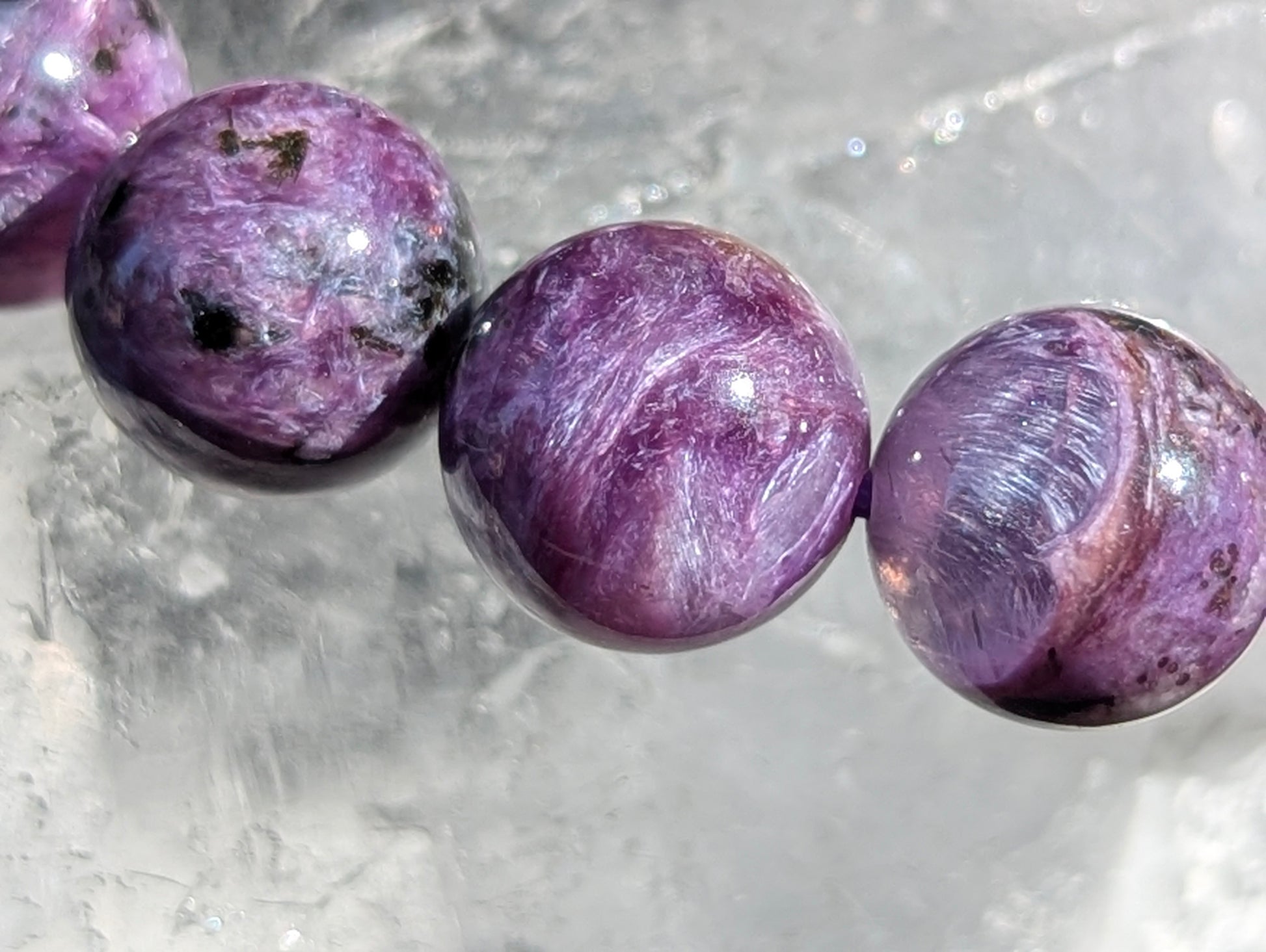 Macro close-up of three AAA Siberian Charoite beads showing intense purple chatoyancy, fibrous swirling patterns, and black aegirine inclusions resting on quartz.