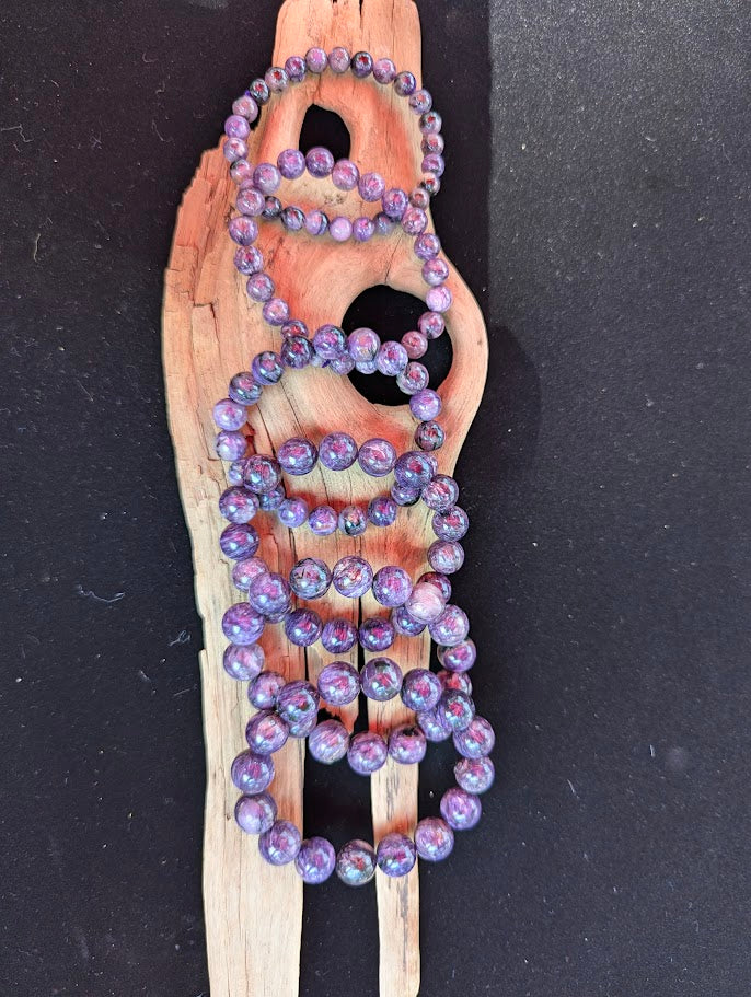 Suite of intuitively selected AAA grade Siberian Charoite beaded stretch bracelets displayed side-by-side on driftwood, showing varied sizes from 7mm to 13mm.