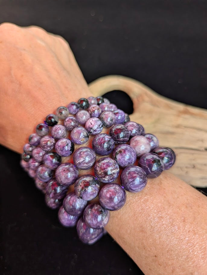 Stack of intuitively selected AAA grade Siberian Charoite beaded bracelets worn together on a wrist, demonstrating size progression from 7mm to 13mm alongside natural black Aegirine and reddish Tinaksite inclusions.