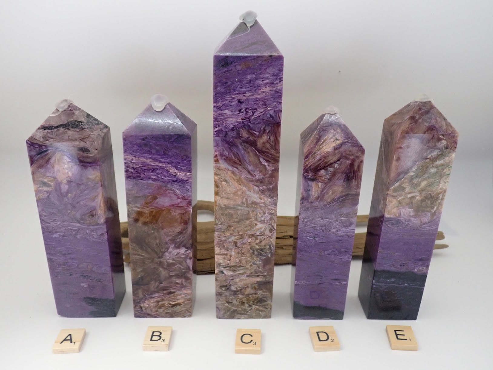 Five polished Charoite towers labeled A through E standing in a row. The set displays a range of quality from deep gemmy violet to golden landscape matrix.