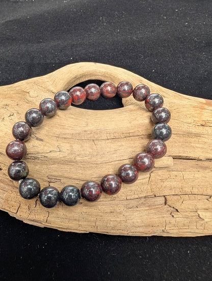 8mm polished Chicken Blood Jasper beaded stretch bracelet resting on a driftwood display, showing dark grey and deep red mottled patterns.