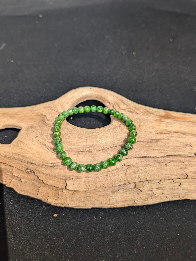 A 4mm Chrome Diopside beaded bracelet resting on natural driftwood, displaying vibrant, translucent emerald-green colors.