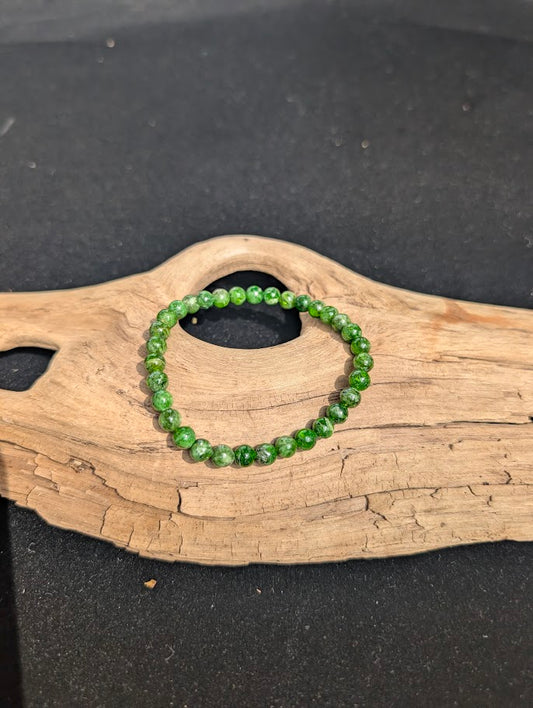 A 4mm Chrome Diopside beaded bracelet resting on natural driftwood, displaying vibrant, translucent emerald-green colors.