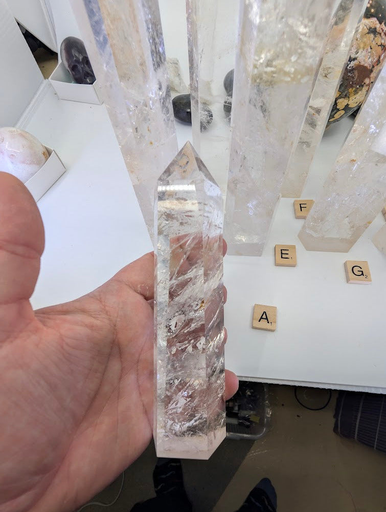 Hand holding a 16cm polished Clear Quartz tower (CQT-A) demonstrating its high transparency and dense internal veils.
