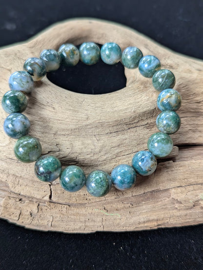 10mm High-Quality Dark Moss Agate gemstone bracelet displayed on textured driftwood highlighting bold green mineral inclusions.