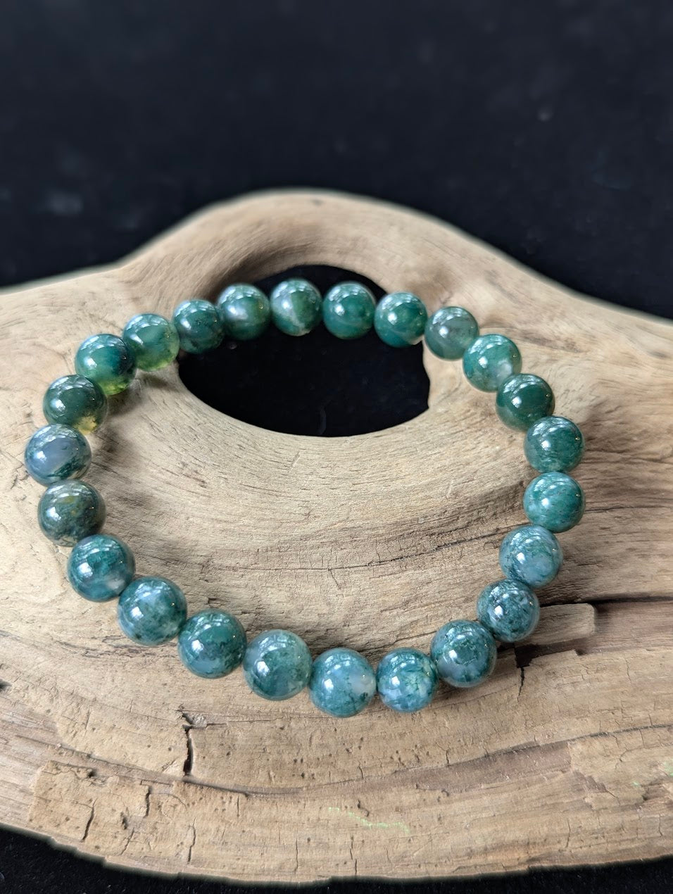9mm High-Quality Dark Moss Agate gemstone bracelet resting on organic driftwood against a dark gallery background.