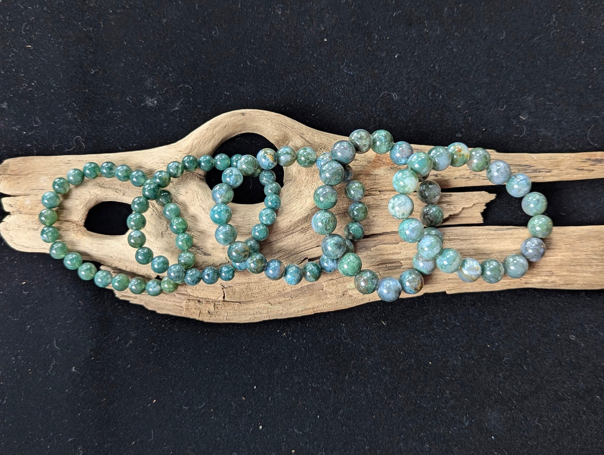 Full lineup of Dark Moss Agate gemstone bracelets across all sizing variants arranged side-by-side on natural driftwood.