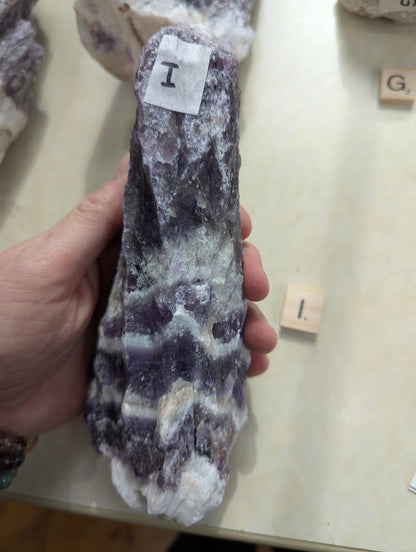 Raw Dream Amethyst specimen held in hand, showing prominent V-shaped chevron patterns across the natural surface.