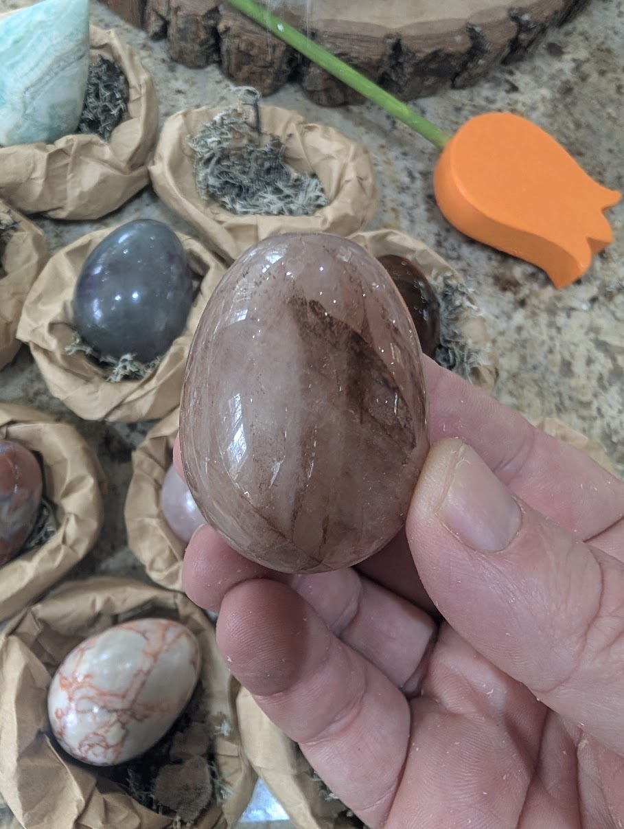 A 2-inch polished Fire Quartz egg held in a hand, displaying a clear to semi-translucent natural quartz base with striking reddish-brown hematite ribbons and inclusions.