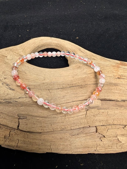4mm polished Fire Quartz beaded stretch bracelet resting on a driftwood display, showing clear quartz with red hematite inclusions.