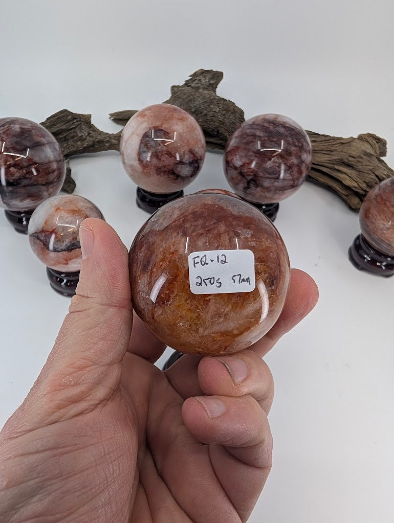 Collector hand scale verification photo of Brazilian Fire Quartz sphere FQ-12, showing the tracking label marked 250 grams and 57mm.